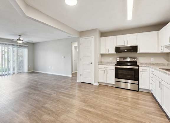 Open-concept apartment interior featuring kitchen with white cabinets and stainless steel oven, flowing into a spacious living area with wood-style flooring.