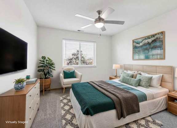 Virtually staged apartment bedroom with ceiling fan, large windows, neutral finishes, and cozy seating area beside the bed.