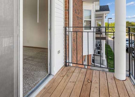 Covered apartment balcony with wood decking, a sliding glass door, brick accents, white support columns, and views of the surrounding apartment community.