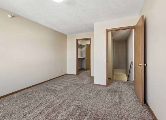 Spacious apartment bedroom with neutral carpet, beige walls, wood-trimmed doors, and an open layout leading to the hallway and closet area.