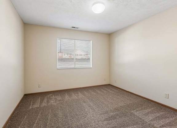 Bright, empty apartment bedroom with neutral carpet, beige walls, wood trim, and a window with blinds that brings in natural light.