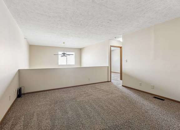 Spacious loft area in an apartment with neutral carpet, a half wall overlooking the lower level, a window for natural light, and an open doorway leading to the hallway.