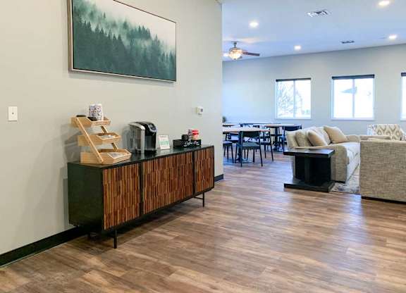 a clubhouse kitchen and tv lounge with wooden floors and furniture, a painting on the wall, and a wooden cabinet with items on top