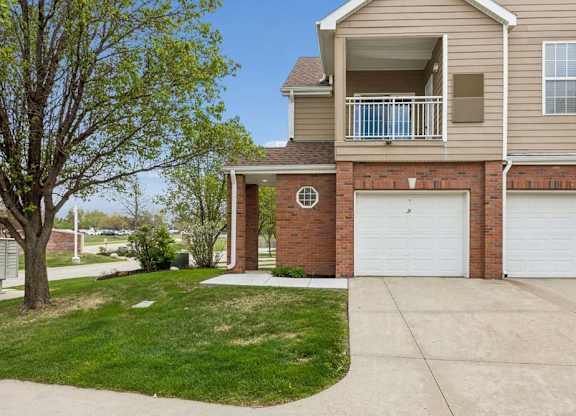 Apartment exterior with brick accents, tan siding, attached garages, balcony, driveway, and grassy lawn.