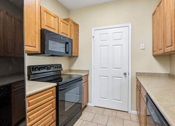Galley-style apartment kitchen with wood cabinets, black appliances, oven, microwave, and tile floor.