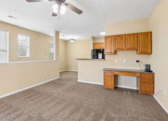 Open apartment living area with carpet, built-in desk cabinetry, wood cabinets, and windows with blinds.