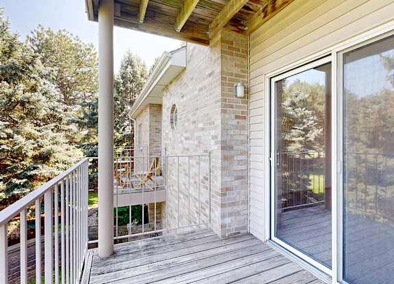 Private apartment balcony with sliding glass door, brick exterior, wood ceiling, and views of surrounding trees and neighboring buildings.