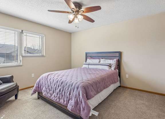 Bright apartment bedroom with neutral carpet, large corner windows, ceiling fan, and a queen-size bed, offering a comfortable and airy interior.