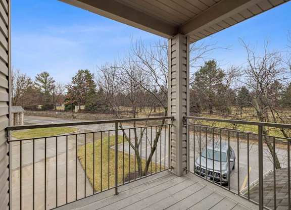 Private apartment balcony with wood decking and metal railing overlooking a parking area and landscaped grounds with mature trees.