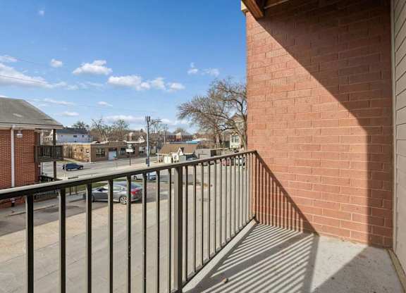 Private apartment balcony with a black metal railing, brick exterior, and neighborhood views under a clear blue sky.