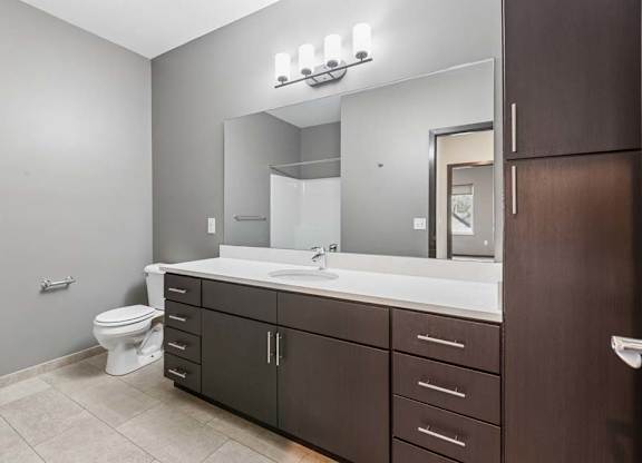 Modern apartment bathroom with a double-sink vanity, dark wood cabinetry, a large mirror, and bright vanity lighting.