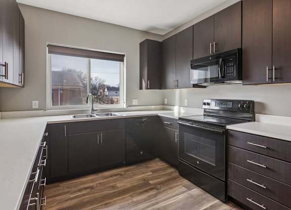 Modern apartment kitchen with dark wood cabinetry, black appliances, white countertops, and a window over the sink.