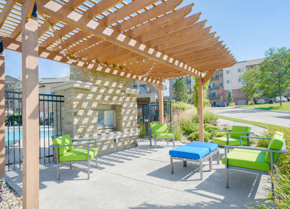 a pergola with green chairs and a blue ottoman in front of a stone fireplace