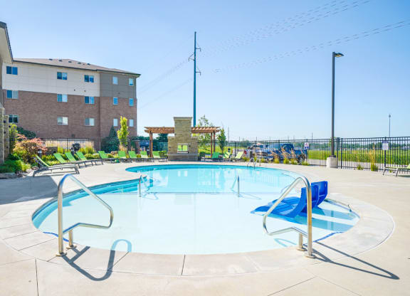 a swimming pool with blue chairs in shallow water and an apartment building in the background