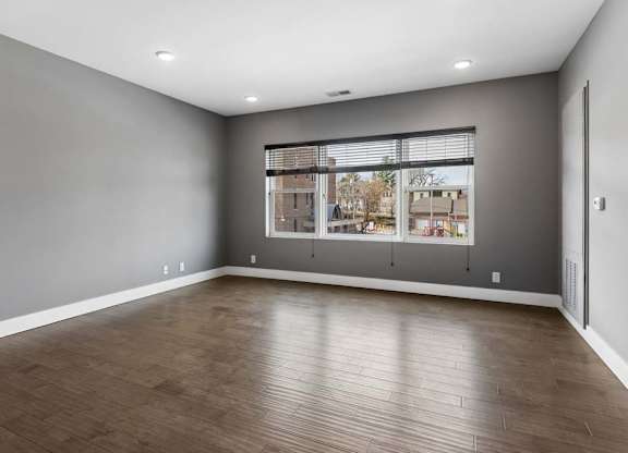 Expansive apartment living room features wood-look flooring, gray walls, and a broad window overlooking the surrounding community.