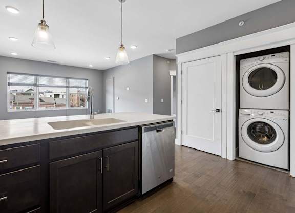 Stacked in-home laundry sits just off the kitchen, beside a dark cabinet island with a deep sink, dishwasher, and pendant lights.