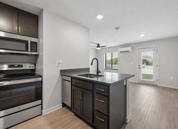 Open-concept apartment kitchen with dark cabinetry, stainless steel appliances, a center island with sink, and a spacious living area with a glass patio door.