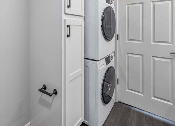 Apartment laundry area with stacked washer and dryer, storage cabinetry, and wood-style flooring.