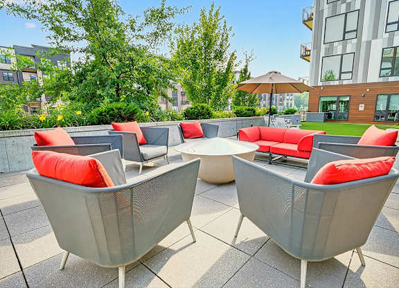 A fire pit surrounded by gray lounge chairs and red-cushions on a patio with nearby greenery.