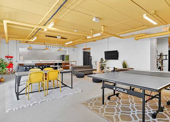 Bright game room with yellow ceiling, featuring a ping pong table, foosball table, and a dining area with four yellow chairs.