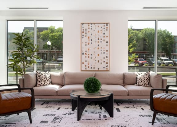 Community lounging space with large beige couch in between two large windows. Two chair sit opposite of the couch with a small coffee table at the center.