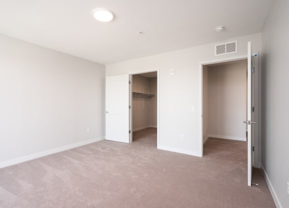 large bedroom with beige carpet and white walls. Two open doors, one leading to the walk in closet and the other leads to the rest of the apartment.