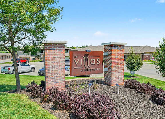 Apartment community entrance sign for The Villas at Falling Waters, set between brick columns with landscaped greenery and parking area in the background.