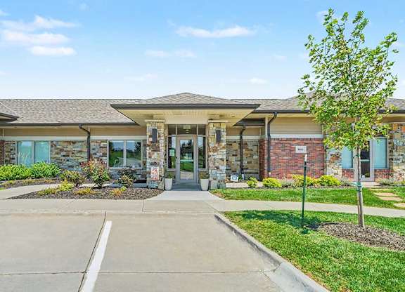 Front view of apartment leasing office with glass double doors, stone and brick exterior, manicured lawn, small tree, and parking spaces in foreground.