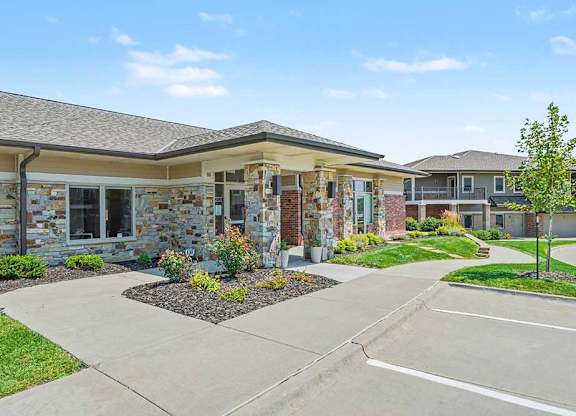 Front view of apartment leasing office with glass double doors, stone and brick exterior, manicured lawn, small tree, and parking spaces in foreground.