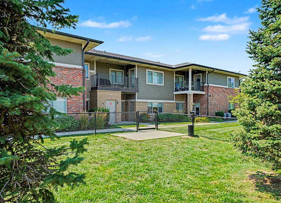 Two-story apartment building with brick and siding exterior, private balconies, and a fenced green lawn area with trees and open sky.