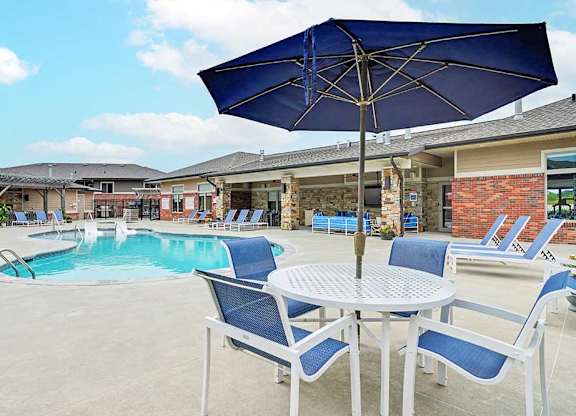 Poolside patio area with round table, blue cushioned chairs, and large umbrella overlooking a resort-style apartment pool and surrounding lounge seating.