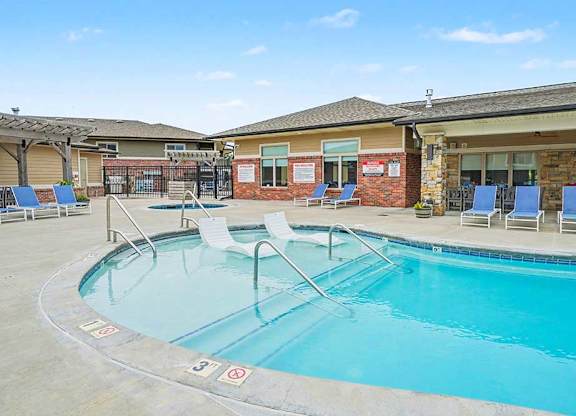 Apartment community pool with clear blue water, metal handrails, and lounge chairs, set beside a covered clubhouse patio with stone and brick exterior.