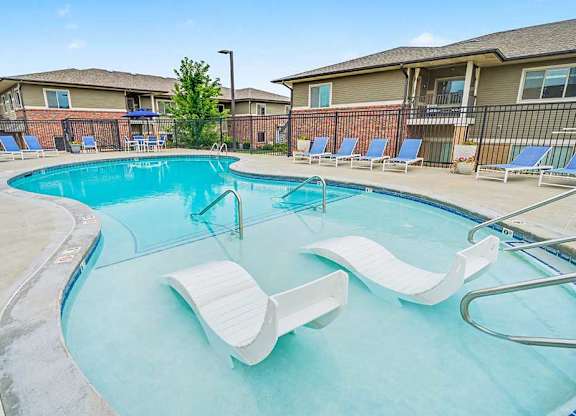 Resort-style apartment pool with curved design, shallow sun shelf and in-water lounge chairs, surrounded by deck seating and fenced residential buildings.