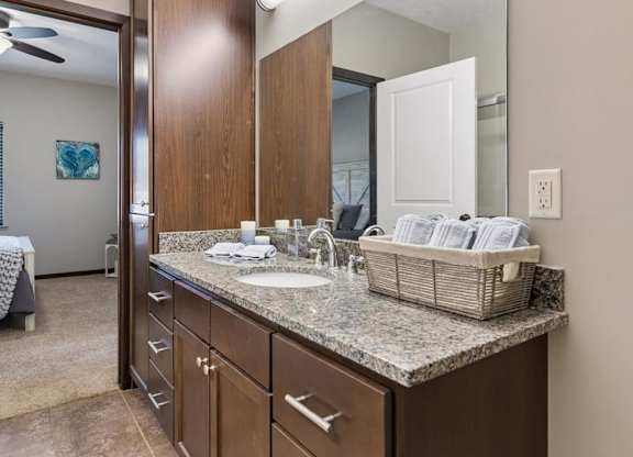 Staged apartment bathroom vanity with speckled countertop, dark wood cabinets, large mirror, and tile flooring.