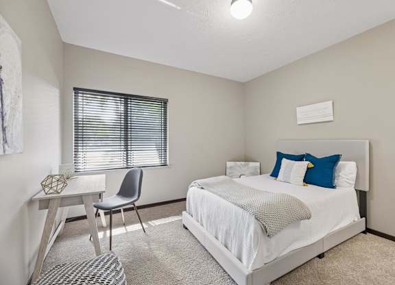 Bright staged apartment bedroom with carpet, upholstered bed, desk, window with blinds, and neutral finishes.