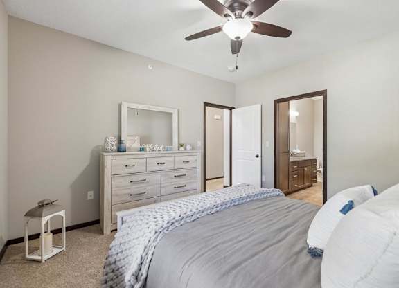 Staged apartment bedroom with ceiling fan, dresser, carpet, and direct access to the bathroom.