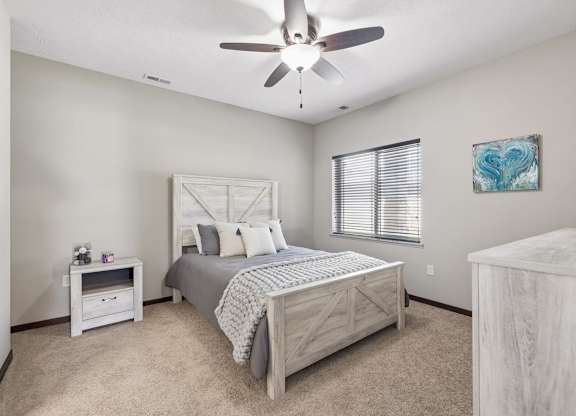 Staged apartment bedroom with ceiling fan, carpet, wood-look bed frame, dresser, and window with blinds.