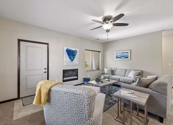 Staged apartment living room with sofa, armchair, ceiling fan, kitchen view, and entry door with tile landing.