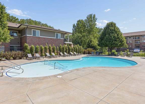 Outdoor apartment community swimming pool with lounge chairs, wide concrete deck, mature trees, and residential buildings.