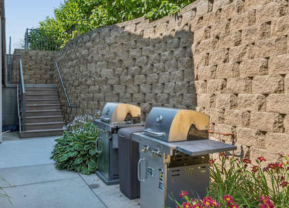 Outdoor grilling amenity at Villas of Omaha at Butler Ridge with two grills, landscaped greenery, and nearby stairs.
