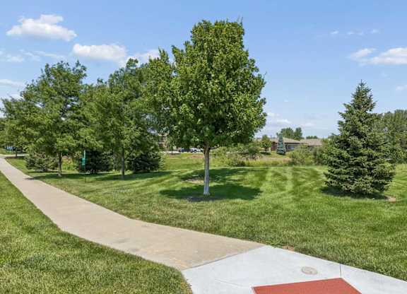 Landscaped walking path at Villas of Omaha at Butler Ridge winding through green lawn and mature trees near apartment buildings.