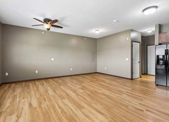 Bright, empty apartment living area with wood-look flooring, neutral walls, ceiling fan, and an open view into the kitchen.