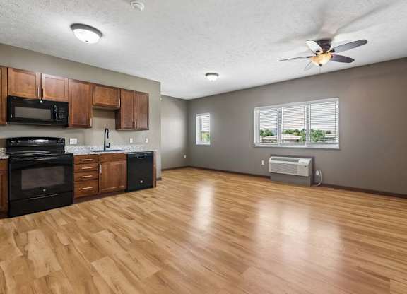 Open-concept apartment kitchen and living area with wood-look flooring, dark appliances, wood cabinetry, ceiling fan, and large windows.