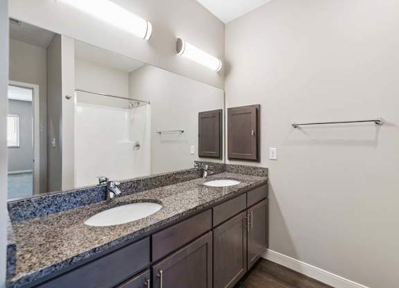 Apartment bathroom with a double-sink vanity, dark cabinetry, granite-look countertops, a large mirror, and modern lighting with towel bars along the wall.