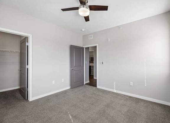 Spacious apartment bedroom with carpeted flooring, a ceiling fan, a walk-in closet, and a window with blinds providing natural light.