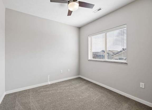 Bright apartment bedroom with carpeted flooring, a ceiling fan, neutral walls, and a window with blinds providing natural light.