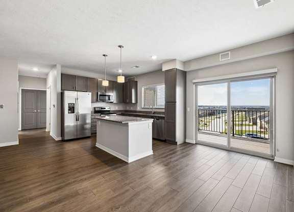 Spacious open-concept apartment living area with wood-style flooring, a modern kitchen featuring a center island, pendant lighting, stainless steel appliances, and a large sliding glass door leading to a private balcony.