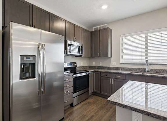 Modern apartment kitchen with dark cabinetry, granite-look countertops, stainless steel appliances, and a large window above the sink providing natural light.