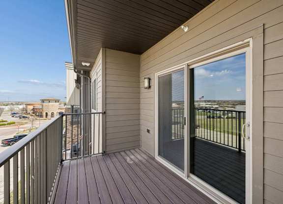 Covered apartment balcony with wood-style decking, metal railings, and a large sliding glass door overlooking the surrounding community.