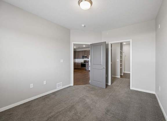 Spacious apartment bedroom with carpeted flooring, neutral walls, and an open layout featuring a walk-in closet and hallway access through gray interior doors.
